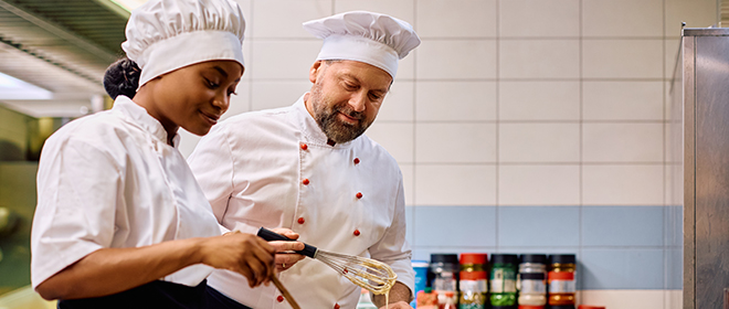 a black female baker apprentice working in a kitchen while a male baker journeyperson supervises